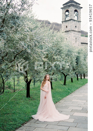 Bride in a pink dress stands near a green olive tree in a grove against the backdrop of a chapel Bride in a pink dress stands near a green olive tree in a grove against the backdrop of a chapel 93419657