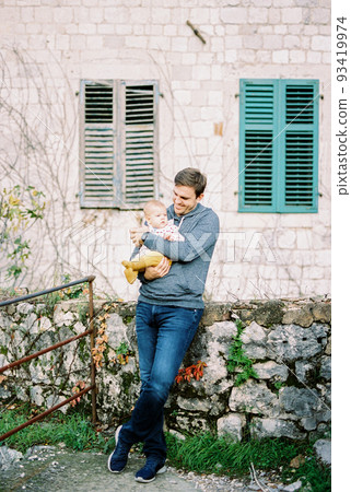 Dad with a baby in his arms stands at the stone wall of the house leaning on the foundation 93419974