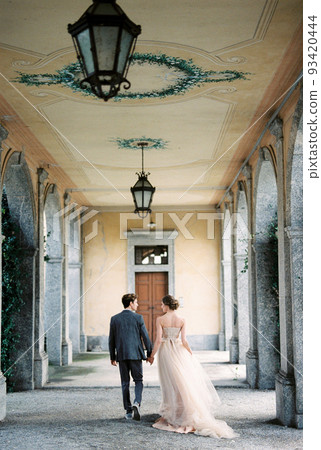 Bride and groom walk holding hands along the terrace of an old villa. Back view 93420444