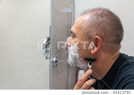 Handsome white-haired beared man shaving off his beard looking in a mirror. Concept of changing style Handsome white-haired beared man shaving off his beard looking in a mirror. Concept of changing style 93423795