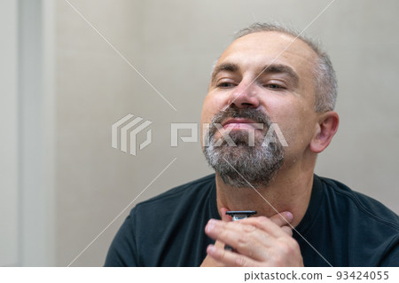 Mature gray-haired man restyling his beard himself at home using razor Mature gray-haired man restyling his beard himself at home using razor 93424055