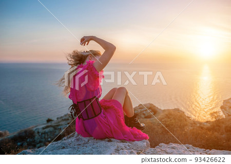 Beautiful young caucasian woman with curly blond hair and freckles. Cute redhead woman portrait in a pink long dress posing on a volcanic rock high above the sea during sunset. 93426622