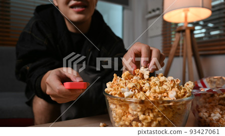 Cropped shot of man eating popcorn and watching television in cozy living room at night. Leisure activity, relaxation, hobby concept 93427061