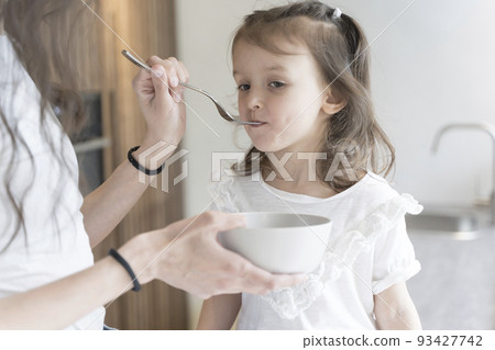 Mom feeds her daughter breakfast in the home kitchen in the morning. Baby food, family, care concept. Mom feeds her daughter breakfast in the home kitchen in the morning. Baby food, family, care concept. 93427742