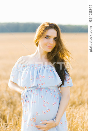 A pregnant woman in a blue dress stands in a field of rye at sunset and holds her hands on her stomach A pregnant woman in a blue dress stands in a field of rye at sunset and holds her hands on her stomach 93428845