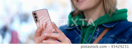 Young woman holding pink mobile phone in park and smiling closeup Young woman holding pink mobile phone in park and smiling closeup 93429231