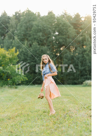 A beautiful young woman holds summer shoes in her hands and walks barefoot on the grass on a summer day 93429751