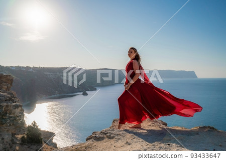 A woman in a red flying dress fluttering in the wind, against the backdrop of the sea. A woman in a red flying dress fluttering in the wind, against the backdrop of the sea. 93433647