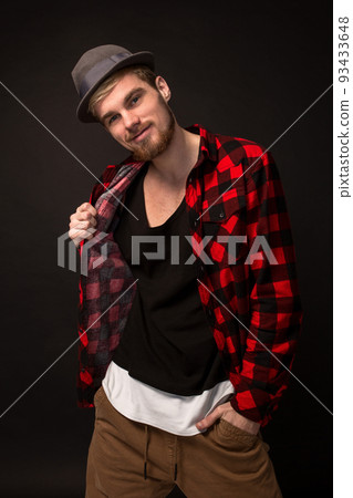 Handsome trendy young guy at the studio on black background. He wears beard and a plaid shirt 93433648