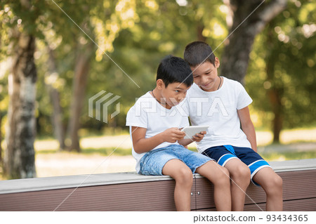 Two boys using together a digital tablet sitting outdoor in park in summer, a child throw an expressive look, selective focus. 93434365