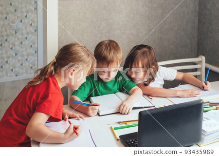 Children sit at a table with pens and notebooks and use a laptop. Home education concept, learning online 93435989