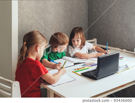 Children sit at a table with pens and notebooks and use a laptop. Homeschooling concept, learning online 93436000