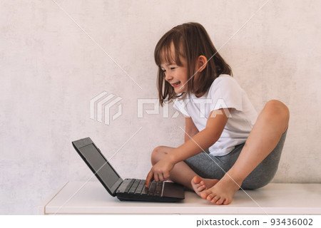 Happy girl sits on the closet and uses a laptop. Happy child is learning online, playing or chatting with friends. 93436002