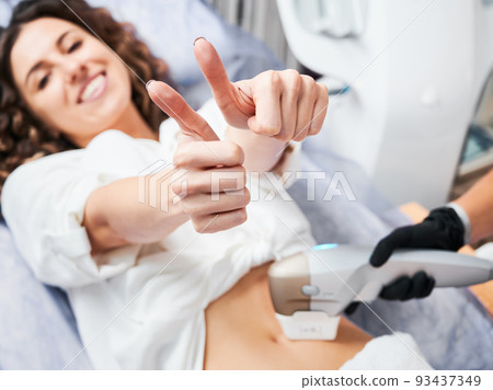 Young satisfied patient with joyful face expression showing thumbs up during SMAS - lifting session in aesthetic medicine clinic. Non-operative method rejuvenation body skin. Focus on female hands. Young satisfied patient with joyful face expression showing thumbs up during SMAS - lifting session in aesthetic medicine clinic. Non-operative method rejuvenation body skin. Focus on female hands. 93437349