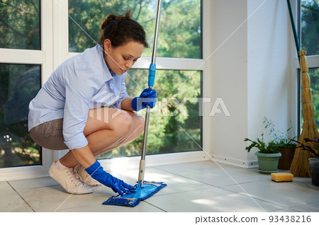 Charming woman, housewife, maid in blue rubber work gloves, putting on a cloth on the mop while mopping the floor in the house veranda. Housekeeping. Professional cleaning service. Chores. Housework Charming woman, housewife, maid in blue rubber work gloves, putting on a cloth on the mop while mopping the floor in the house veranda. Housekeeping. Professional cleaning service. Chores. Housework 93438216