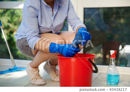 Details: gloved hands of housewife squeezing a floor cleaning cloth into a red bucket during a general cleaning of the house and veranda. Housekeeping. Housework. Chores. Professional cleaning service Details: gloved hands of housewife squeezing a floor cleaning cloth into a red bucket during a general cleaning of the house and veranda. Housekeeping. Housework. Chores. Professional cleaning service 93438217
