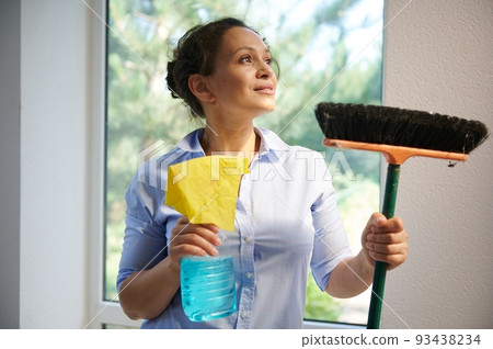 Close-up portrait of a Latin American pretty woman, housewife in blue casual shirt, looking aside, holding a broom and rag with detergent in her hands. Housekeeping and spring cleaning service concept 93438234