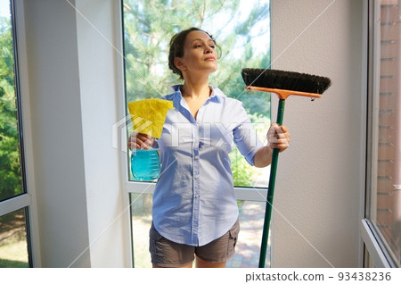 Beautiful woman, housewife, maid, housemaker looking aside, standing in the veranda of a countryside house and holding broom, cloth and detergent. Cleaning, chores, household and housekeeping concept Beautiful woman, housewife, maid, housemaker looking aside, standing in the veranda of a countryside house and holding broom, cloth and detergent. Cleaning, chores, household and housekeeping concept 93438236