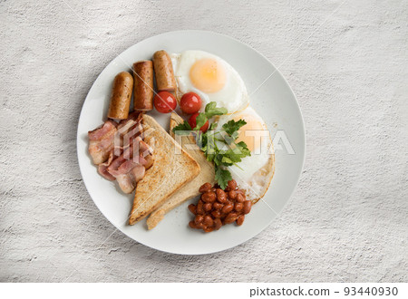 A white plate with scrambled eggs, bacon, beans, sausages, tomatoes and croutons on a light background. 93440930
