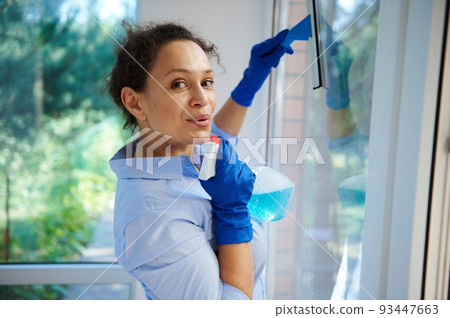 Beautiful multi-ethnic woman, cute housewife happily performing household chores, wiping panoramic veranda windows with detergent spray, removes stains with a glass cleaning and scraper. Housekeeping 93447663