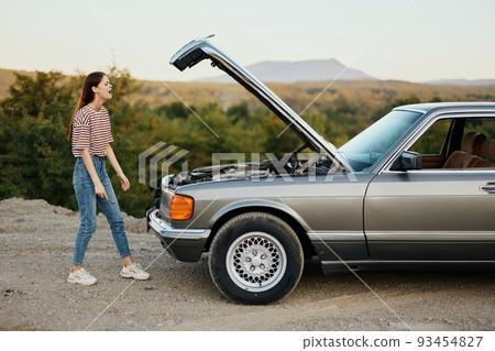 A woman opens the hood of a broken down car and tries to find the cause of the breakdown on the road during a nature trip alone and is sad 93454827