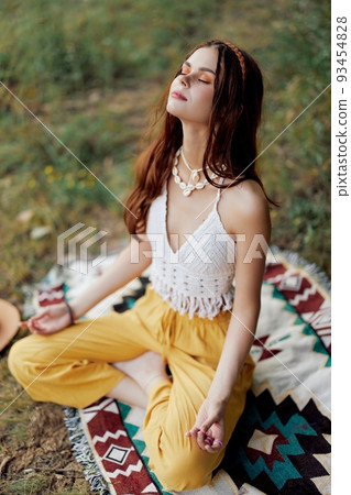 A young hippie woman meditates in nature in the park, sitting in a lotus position on her colorful plaid and enjoying harmony with the world in eco-clothing 93454828