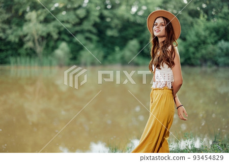 A young woman in a hippie look travels in nature by the lake wearing a hat and yellow pants in the fall 93454829