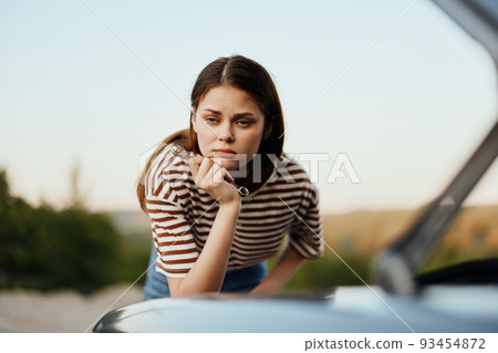 A woman with a wrench looks sadly and sadly under the open hood of her car and can't fix it from a roadside breakdown while traveling alone 93454872