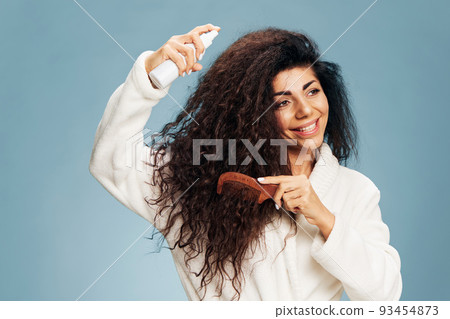 Hairstyling routine. Overjoyed tanned curly Latin lady in bathrobe Spraying On Hair For Repair split ends posing isolated on pastel blue background, using hairbrush, smiling. Copy space offer 93454873