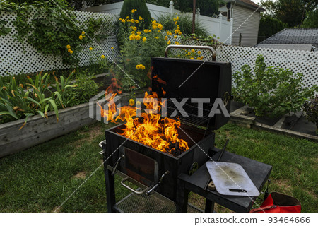 Brightly burning in metal box logs firewood for barbecue outdoor on sunny day. Orange high flame and white smoke on blurred blue sky and green grass background. Camping, safety and tourism concept. Brightly burning in metal box logs firewood for barbecue outdoor on sunny day. Orange high flame and white smoke on blurred blue sky and green grass background. Camping, safety and tourism concept. 93464666