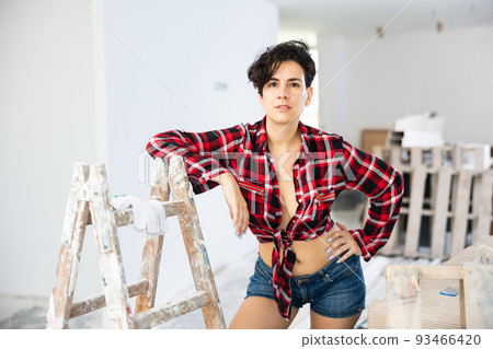 Young brunette in checkered shirt and shorts standing in house being renovated Young brunette in checkered shirt and shorts standing in house being renovated 93466420