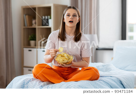 happy girl eating crisps sitting on bed at home 93471184