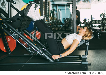 A young woman does exercises for her legs and buttocks while lying on a modern simulator in the gym. A girl does sports in a fitness studio A young woman does exercises for her legs and buttocks while lying on a modern simulator in the gym. A girl does sports in a fitness studio 93476072