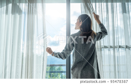 Rear view of happiness Asian woman open curtains for looking beautiful view outside her room. 93478387