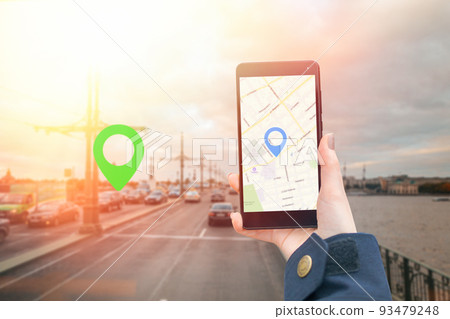A woman's hand in a jacket holds a smartphone with an online map app. In the background is a blurred empty road with icon of location.The concept of Internet navigation and modern technologies A woman's hand in a jacket holds a smartphone with an online map app. In the background is a blurred empty road with icon of location.The concept of Internet navigation and modern technologies 93479248