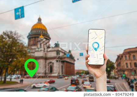 A female's hand holds a smartphone with an online map app. In the background is a city street with building and green location icon.Concept of online navigation and GPS 93479249