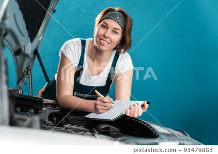 Portrait of young smiling mechanic woman in coveralls signs documents leaning at the car. The concept of woman's equality and works at auto repair shop Portrait of young smiling mechanic woman in coveralls signs documents leaning at the car. The concept of woman's equality and works at auto repair shop 93479883
