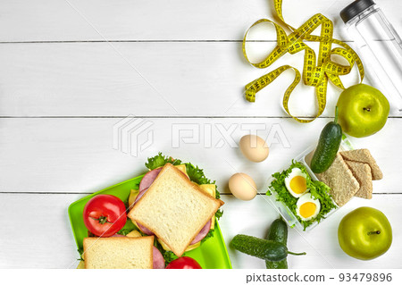 Lunch boxes with sandwiches and fresh vegetables, bottle of water and eggs on white wooden background. Top view with copy space Lunch boxes with sandwiches and fresh vegetables, bottle of water and eggs on white wooden background. Top view with copy space 93479896
