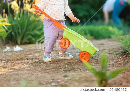 Baby playing with a plastic toy cart at the summer garden. Close up. Concept of gardening and children's education Baby playing with a plastic toy cart at the summer garden. Close up. Concept of gardening and children's education 93480452