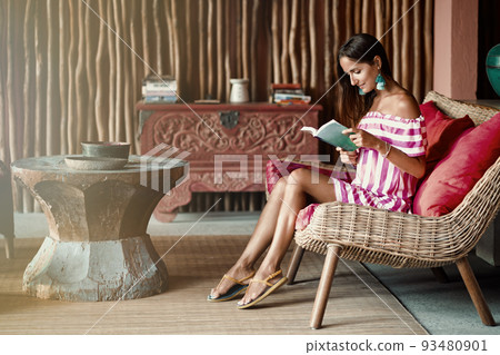 Beautiful brunette woman sitting on a pink couch and reading a book. Interior in ethnic style. Light from the window Beautiful brunette woman sitting on a pink couch and reading a book. Interior in ethnic style. Light from the window 93480901