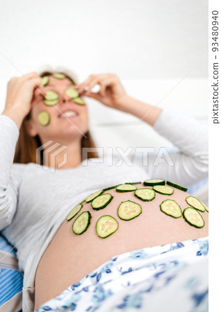 A Caucasian pregnant woman lies on her bed and puts slices of cucumbers on her face and tummy, spa-day. Concept of pregnancy and natural self-care. Close up 93480940