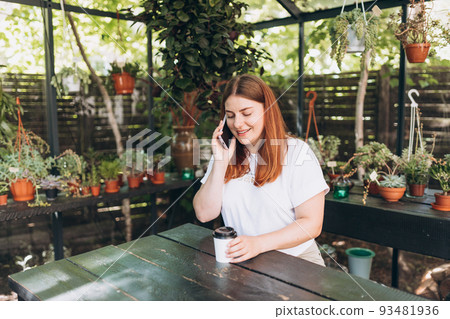 Happy Girl sitting at a table with a coffee using mobile phone. Summer time. Young beautiful woman with coffee paper cup and smartphone while sitting at cafe. Technology banner 93481936