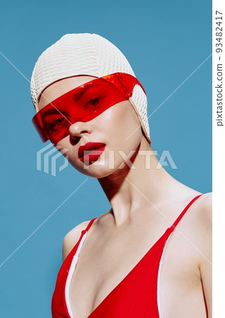 A girl in unusual red glasses and a swimming cap looks into the camera posing on a blue studio background. Art fashion concept for design 93482417