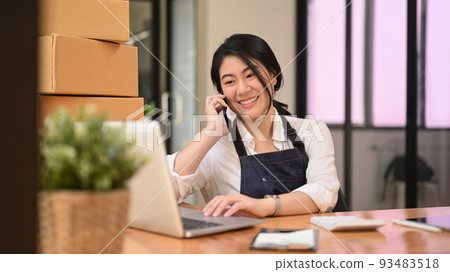 Smiling asian woman online seller using laptop and receiving order on mobile phone. Online selling, e-commerce concept Smiling asian woman online seller using laptop and receiving order on mobile phone. Online selling, e-commerce concept 93483518