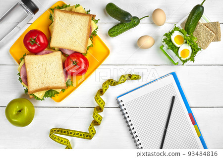Top view planning notebook with copy space and healthy breakfast. Sandwich with ham and cheese, eggs, cucumber, tomato and bottle of water on white wooden table. 93484816