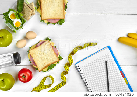 Top view planning notebook with copy space and healthy breakfast. Sandwich with ham and cheese, eggs, cucumber, tomato and bottle of water on white wooden table. Top view planning notebook with copy space and healthy breakfast. Sandwich with ham and cheese, eggs, cucumber, tomato and bottle of water on white wooden table. 93484829