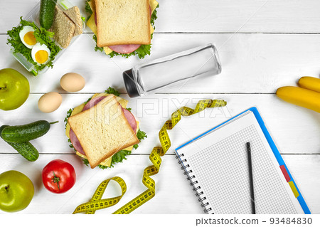 Top view planning notebook with copy space and healthy breakfast. Sandwich with ham and cheese, eggs, cucumber, tomato and bottle of water on white wooden table. Top view planning notebook with copy space and healthy breakfast. Sandwich with ham and cheese, eggs, cucumber, tomato and bottle of water on white wooden table. 93484830