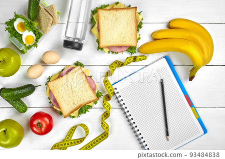 Top view planning notebook with copy space and healthy breakfast. Sandwich with ham and cheese, eggs, cucumber, tomato and bottle of water on white wooden table. 93484838