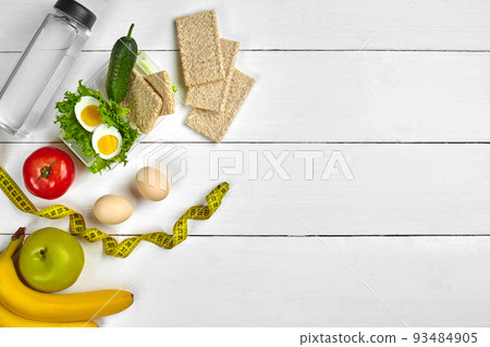 Lunch box with healthy food. Eggs, vegetables, fruits and bottle of water on white wooden background. Top view with copy space Lunch box with healthy food. Eggs, vegetables, fruits and bottle of water on white wooden background. Top view with copy space 93484905