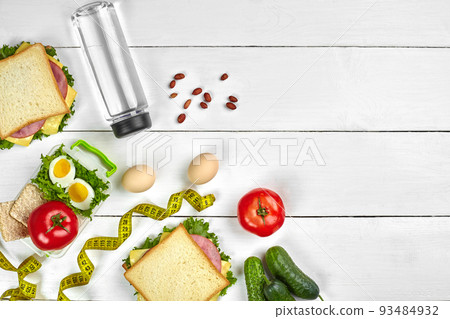 Lunch. Sandwich and fresh vegetables, bottle of water, nuts and fruits on white wooden background. Healthy eating concept. Top view with copy space 93484932